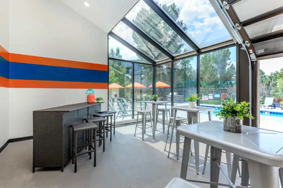 Glass-enclosed community room with a bar and seating, overlooking a pool.