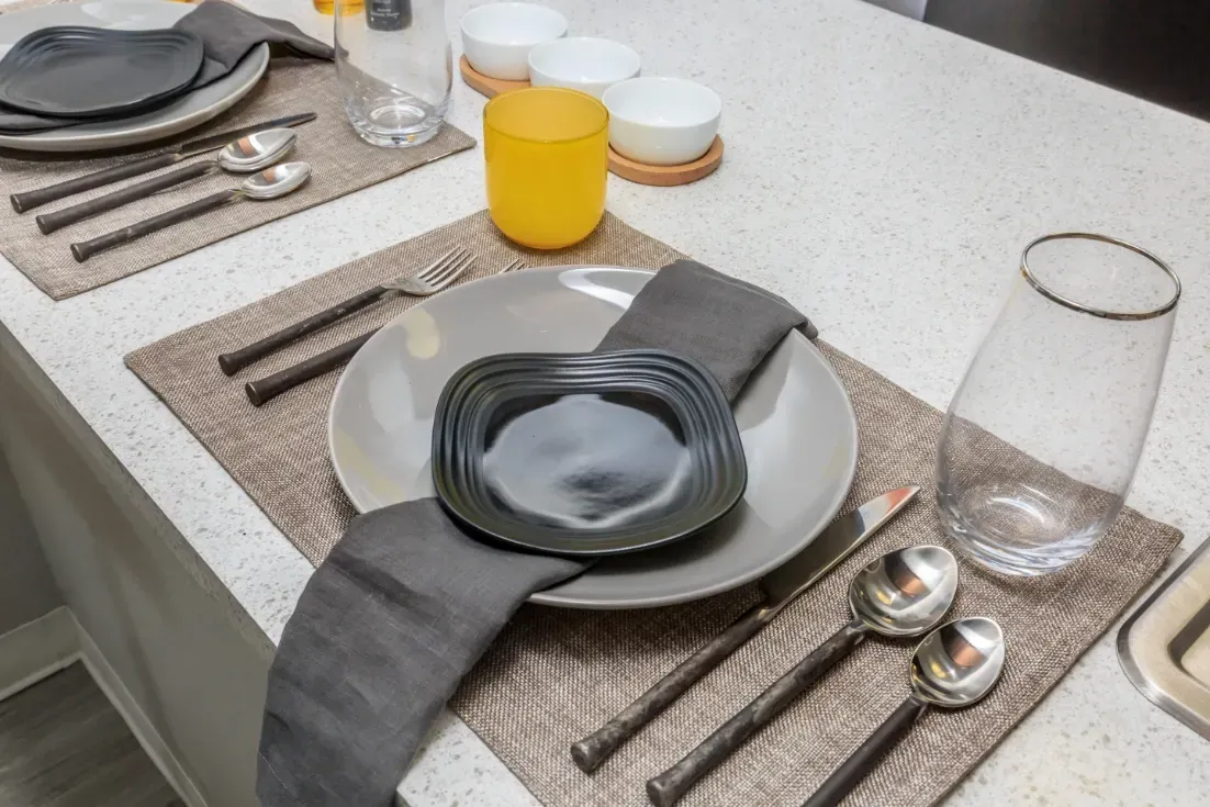 Table setting for two with modern plates, silverware, and glassware on a kitchen counter.