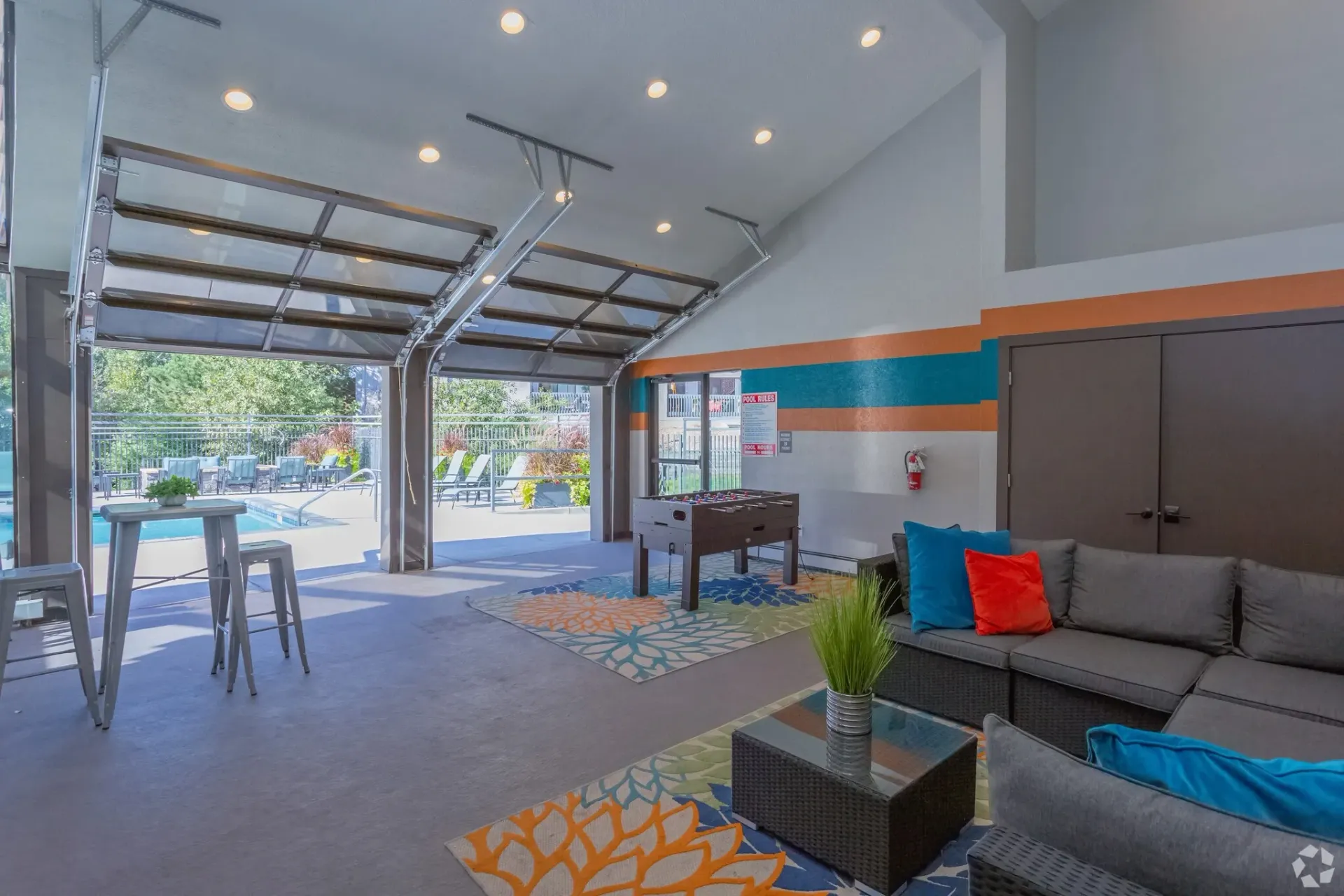 Open garage-style living room with gray sofa, bar stools, and colorful rug, facing an open street view.