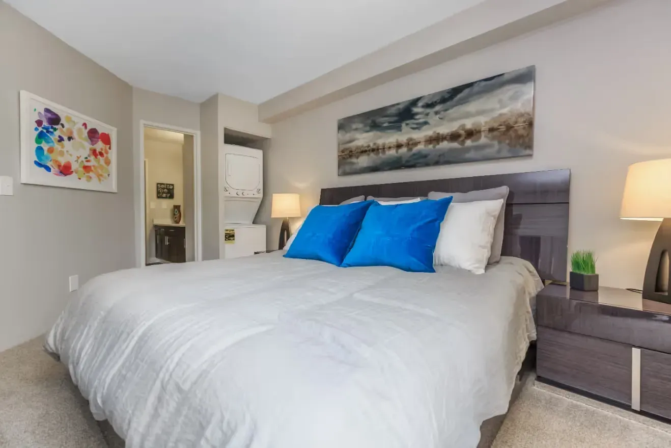 A cozy bedroom featuring a king-size bed with blue and white pillows, a stacked washer and dryer, and modern art.