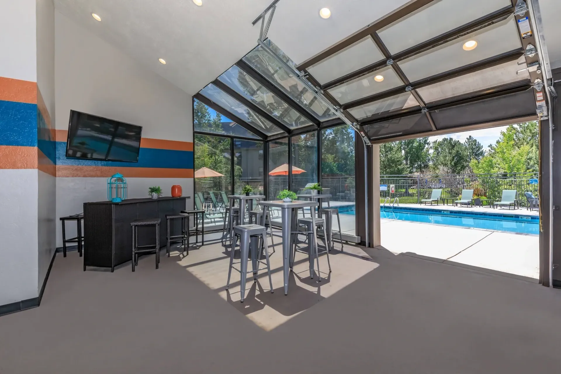 Covered outdoor patio with tables and bar seating beside a pool, with glass roof and open view outside