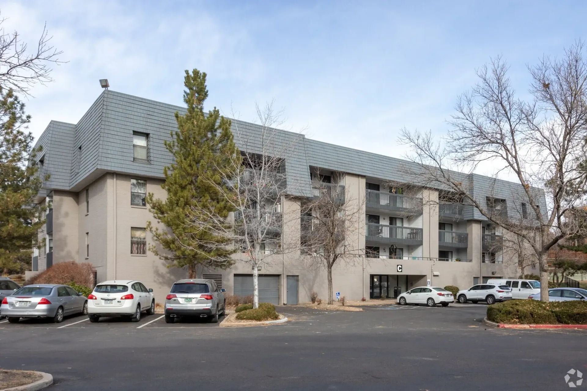 Apartment building with gray roof, parked cars, and leafless trees on a cloudy day