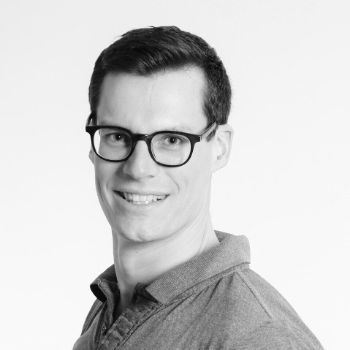 Un homme portant des lunettes et un polo sourit sur une photo en noir et blanc.