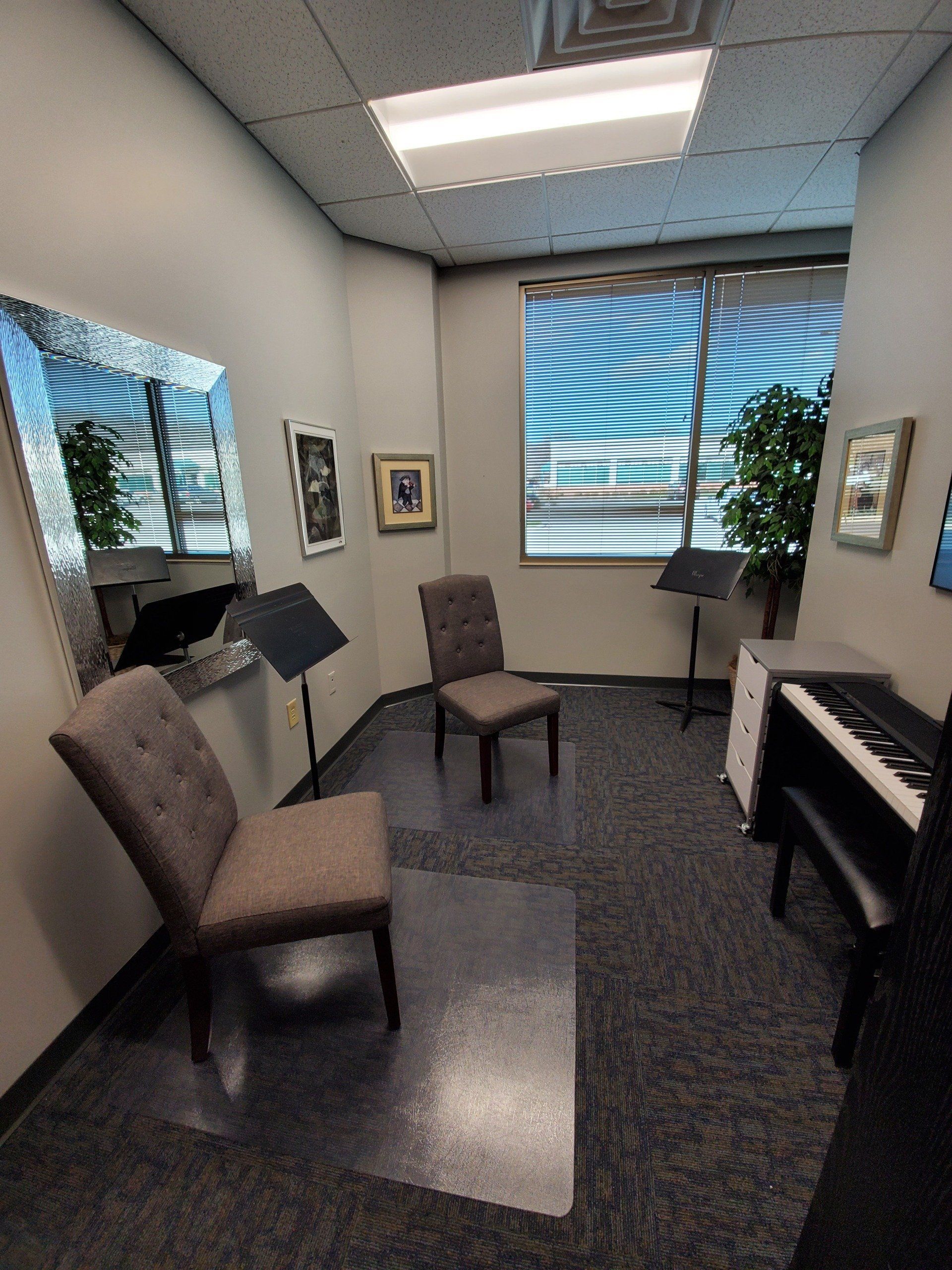 A small music practice room with a piano, chairs, music stands, and a window with a mirrored wall.