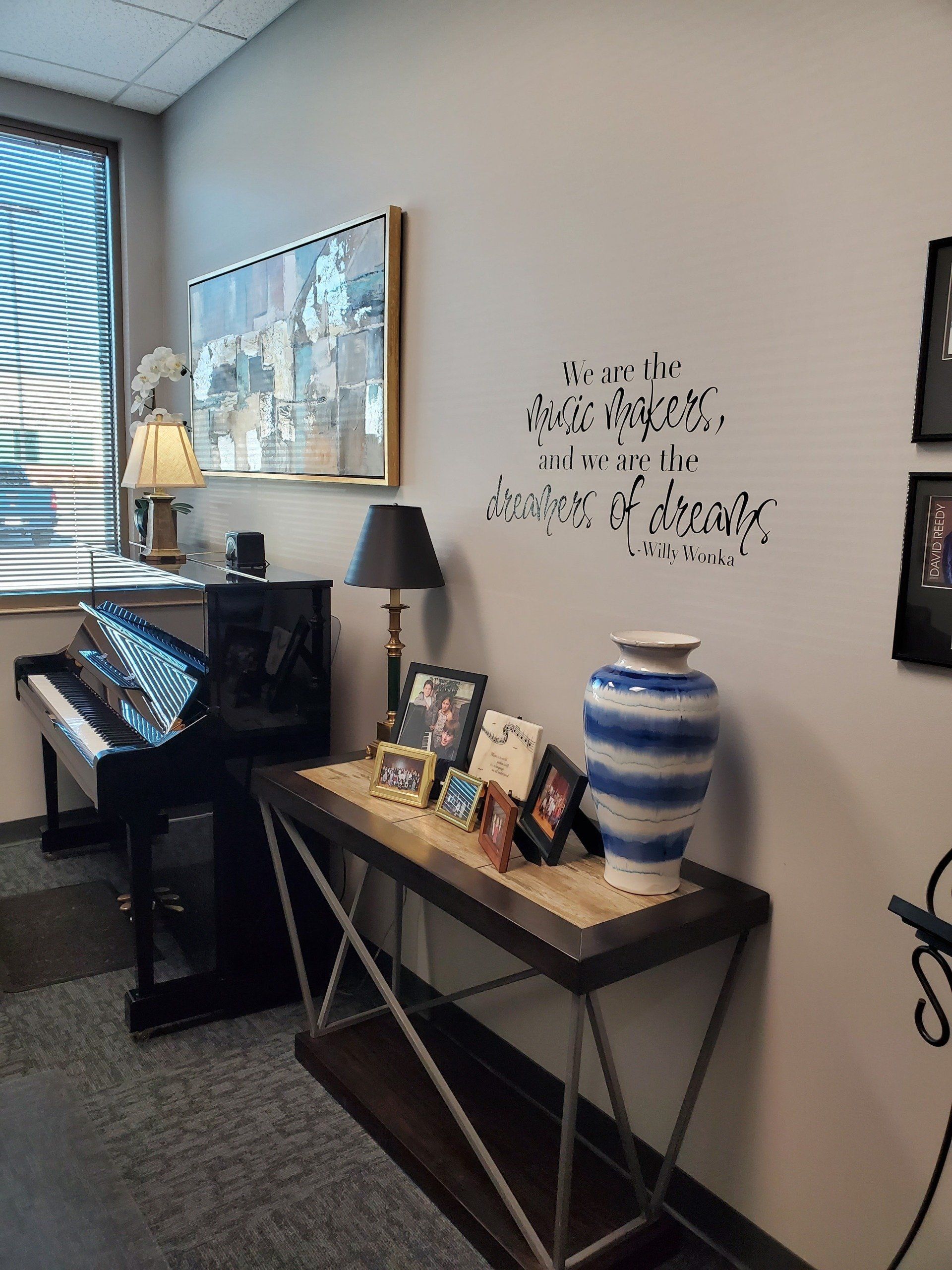 A piano, a table with framed photos, a vase, and a lamp against a gray wall with a window.