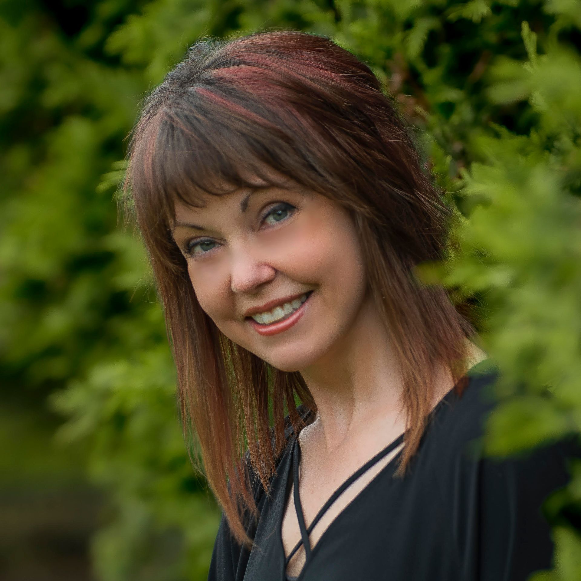 Woman with brown hair and bangs smiles, in front of green foliage.