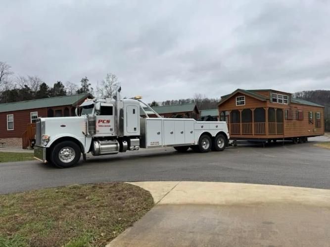 A tow truck hauling a small cabin on a trailer. Cloudy sky, residential setting.