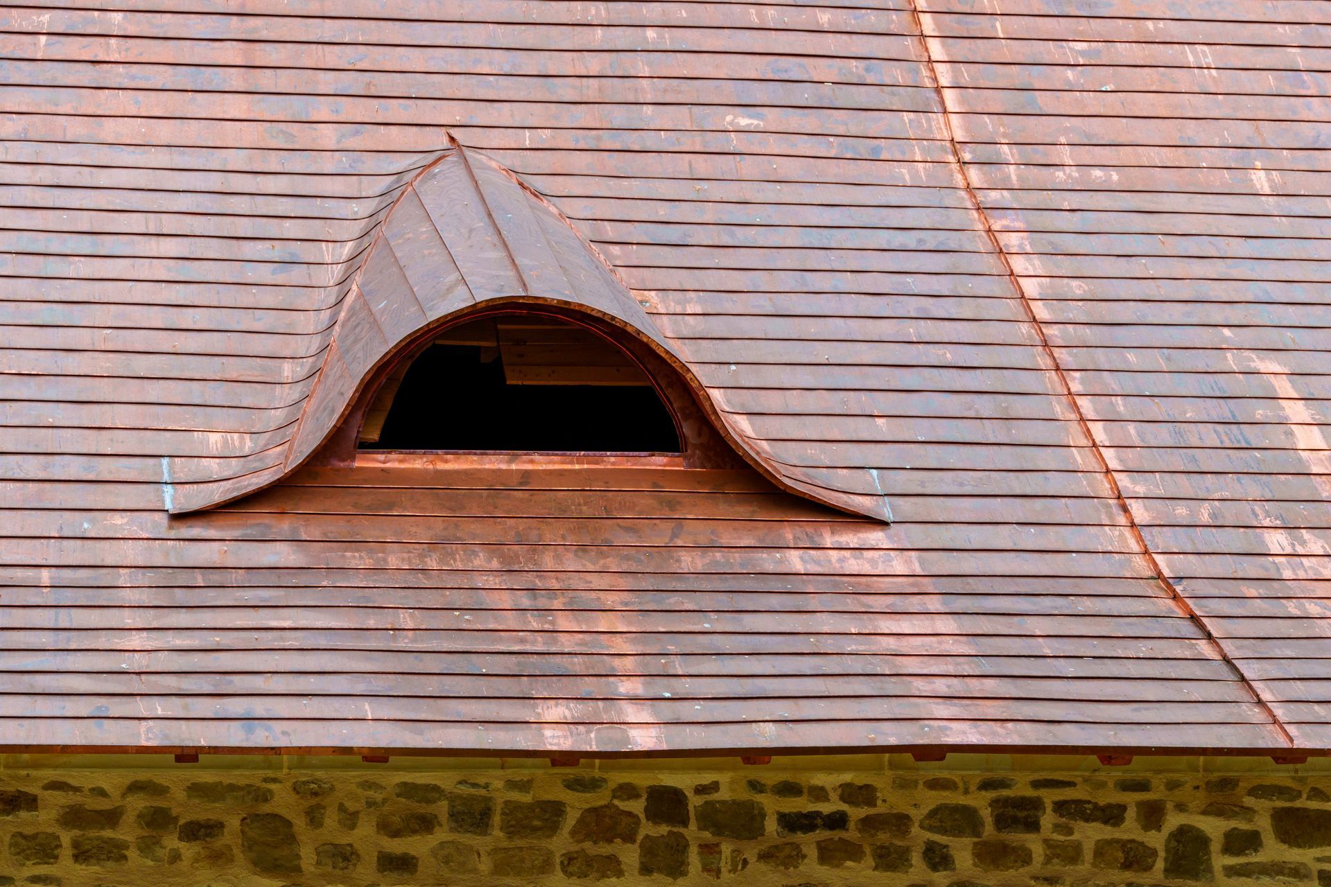 Roof with a small arched dormer opening on reddish shingles above a stone wall