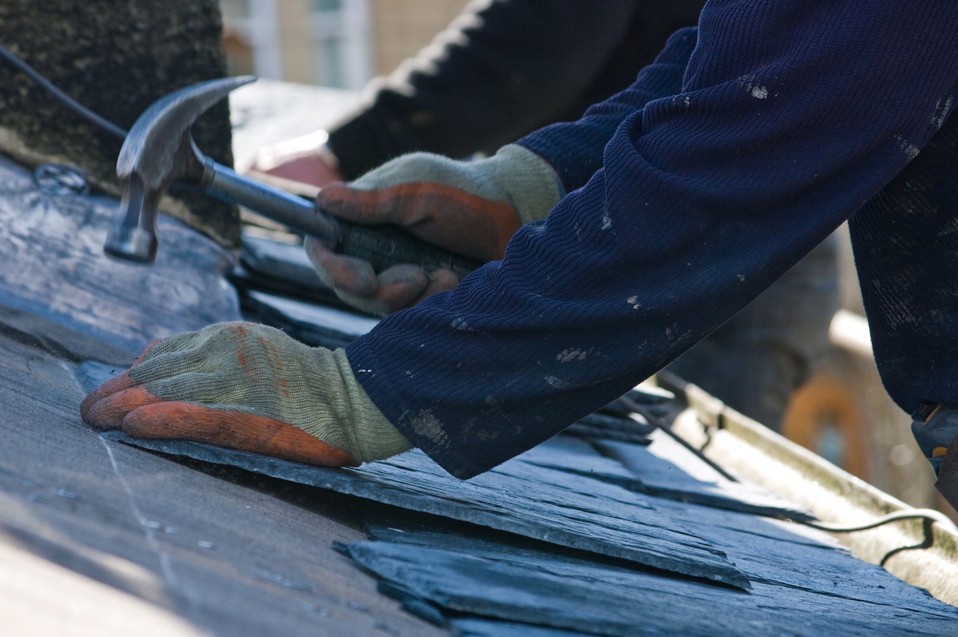 Gloved hands hammering roofing nails into blue shingles on a sloped roof