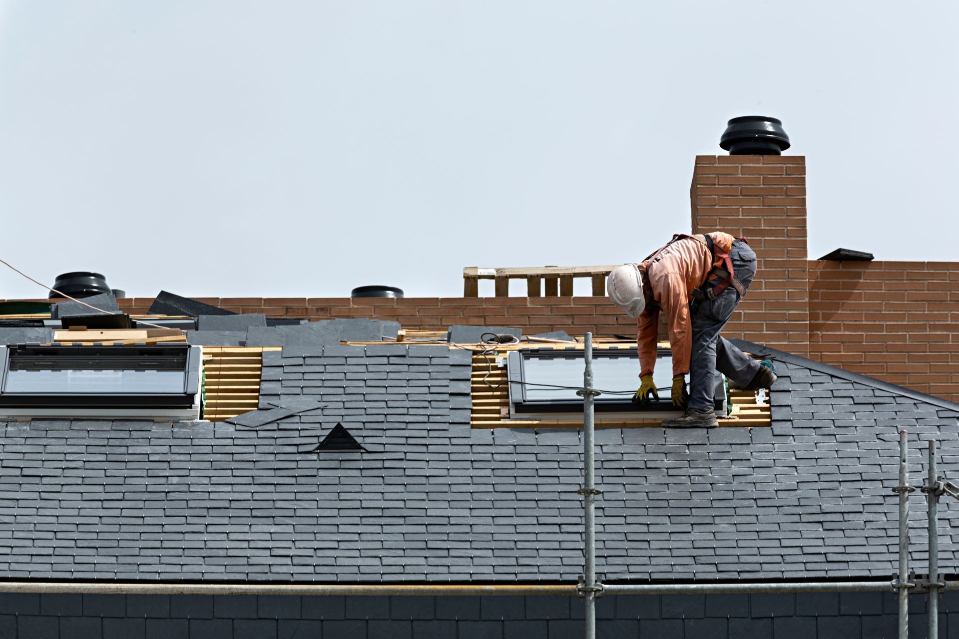 Worker on a gray roof near a chimney, bending over and installing shingles.