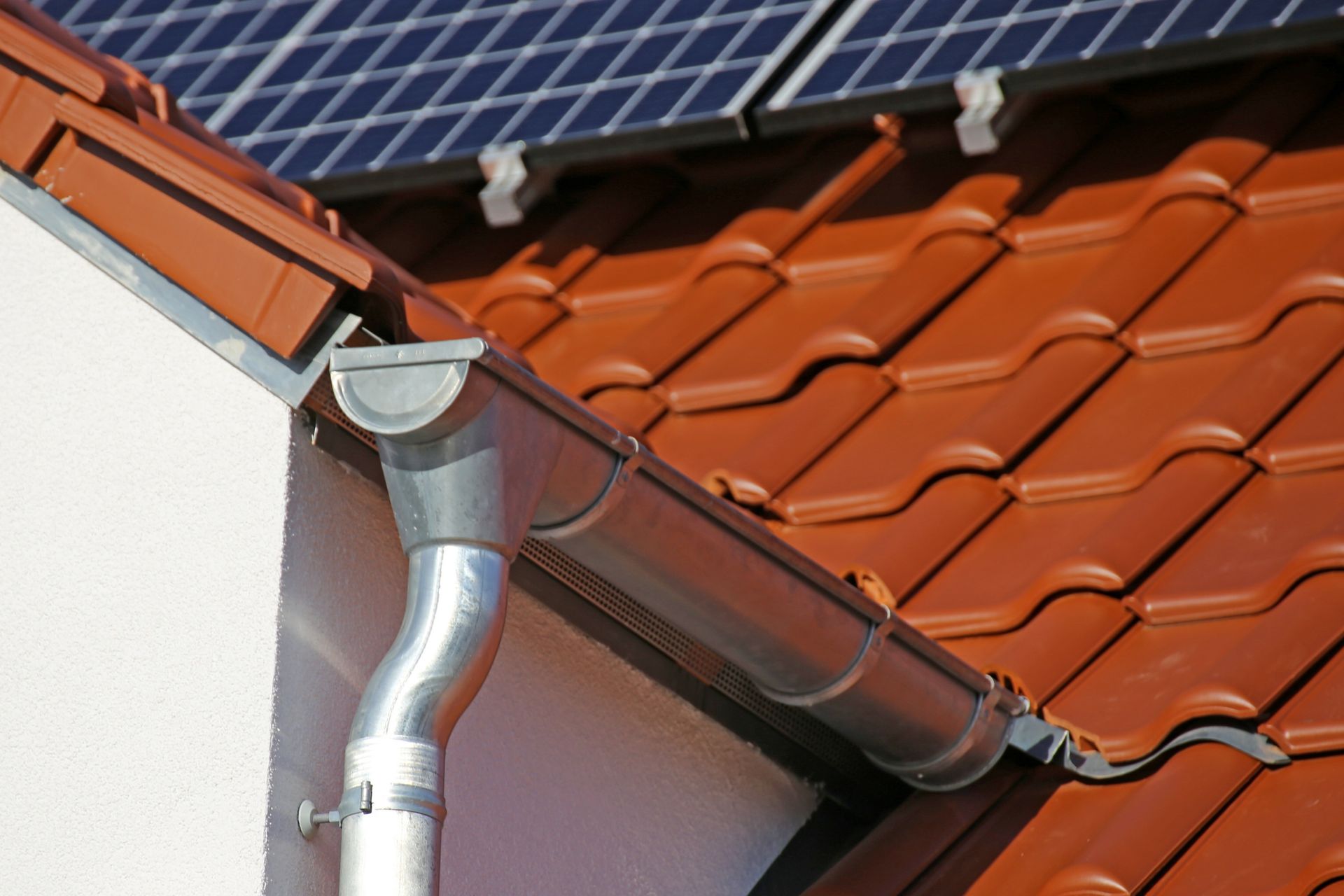 Close-up of a red tiled roof with a rain gutter and downspout beside solar panels