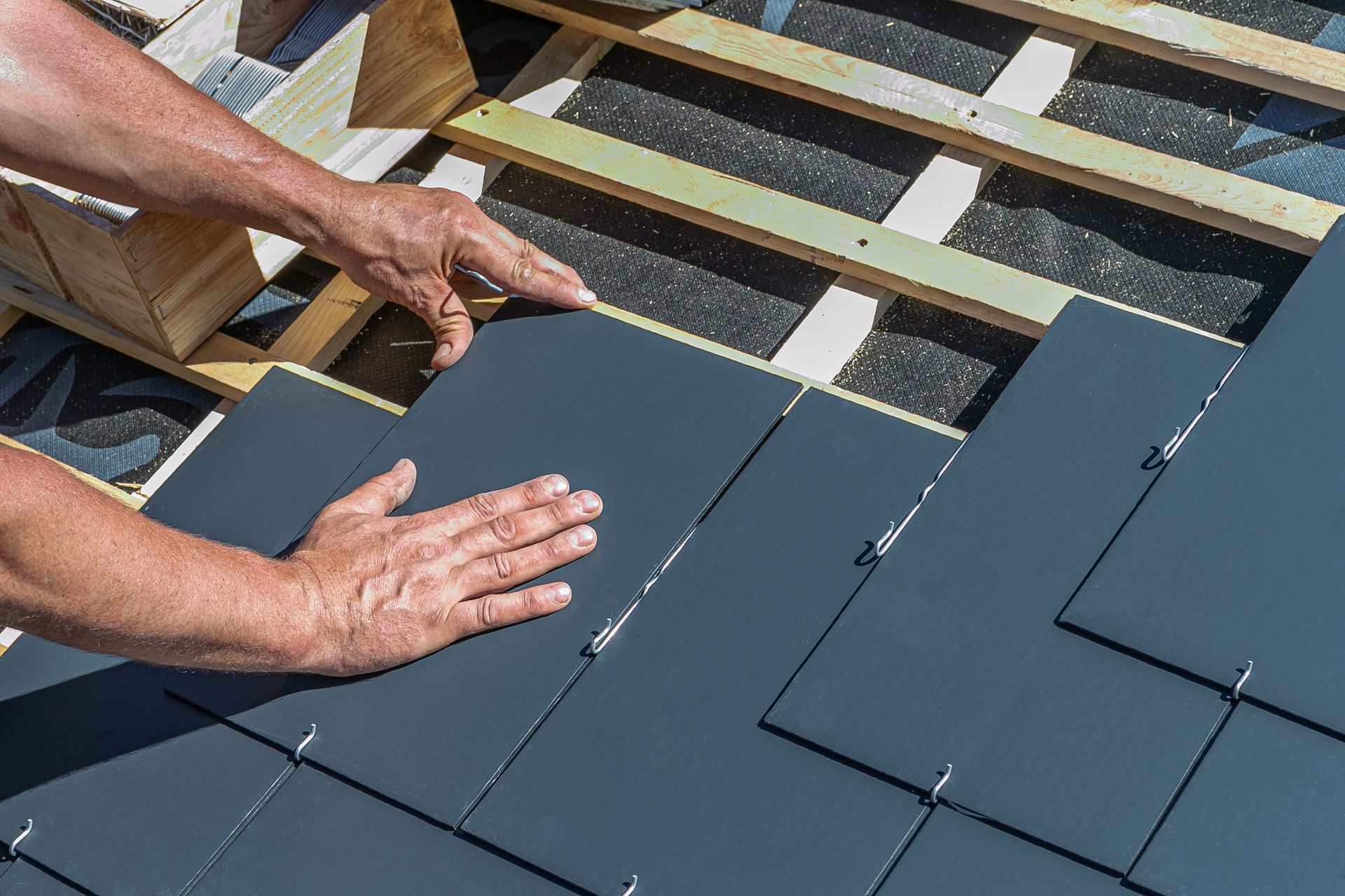 Hands installing dark gray roofing shingles on a roof frame