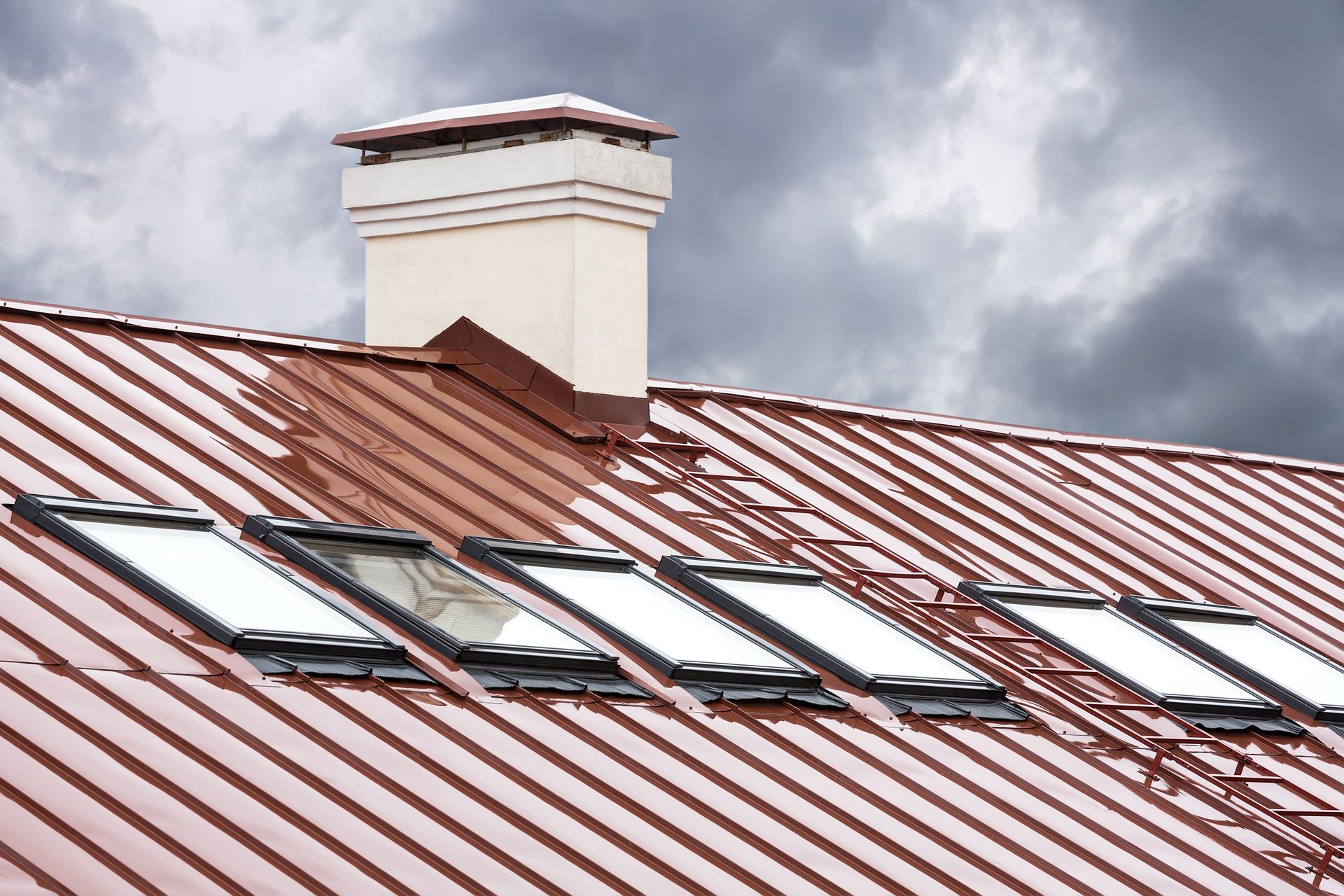 Red metal roof with skylights and a white chimney under cloudy sky