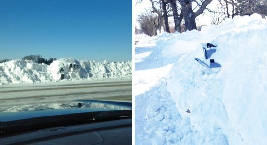 Snow-covered road and mountains seen from a car, with a snowy bank and sign partially buried in snow.