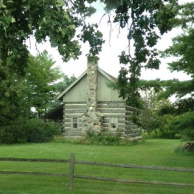 Small stone-and-log cabin in a grassy park, framed by trees and a wooden fence.
