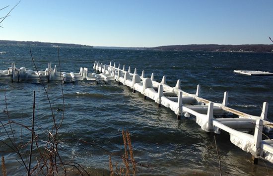 Flooded lakeside walkway with white railings under a cloudy sky, waves crashing along the shore