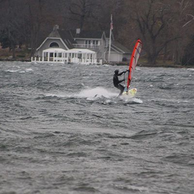 Windsurfer on choppy water near a shoreline house, holding a red sail.