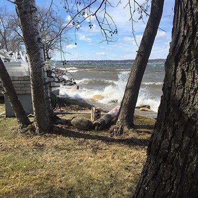 Windy lakeshore with waves crashing against the shore, trees in the foreground, and a person lying near the water.