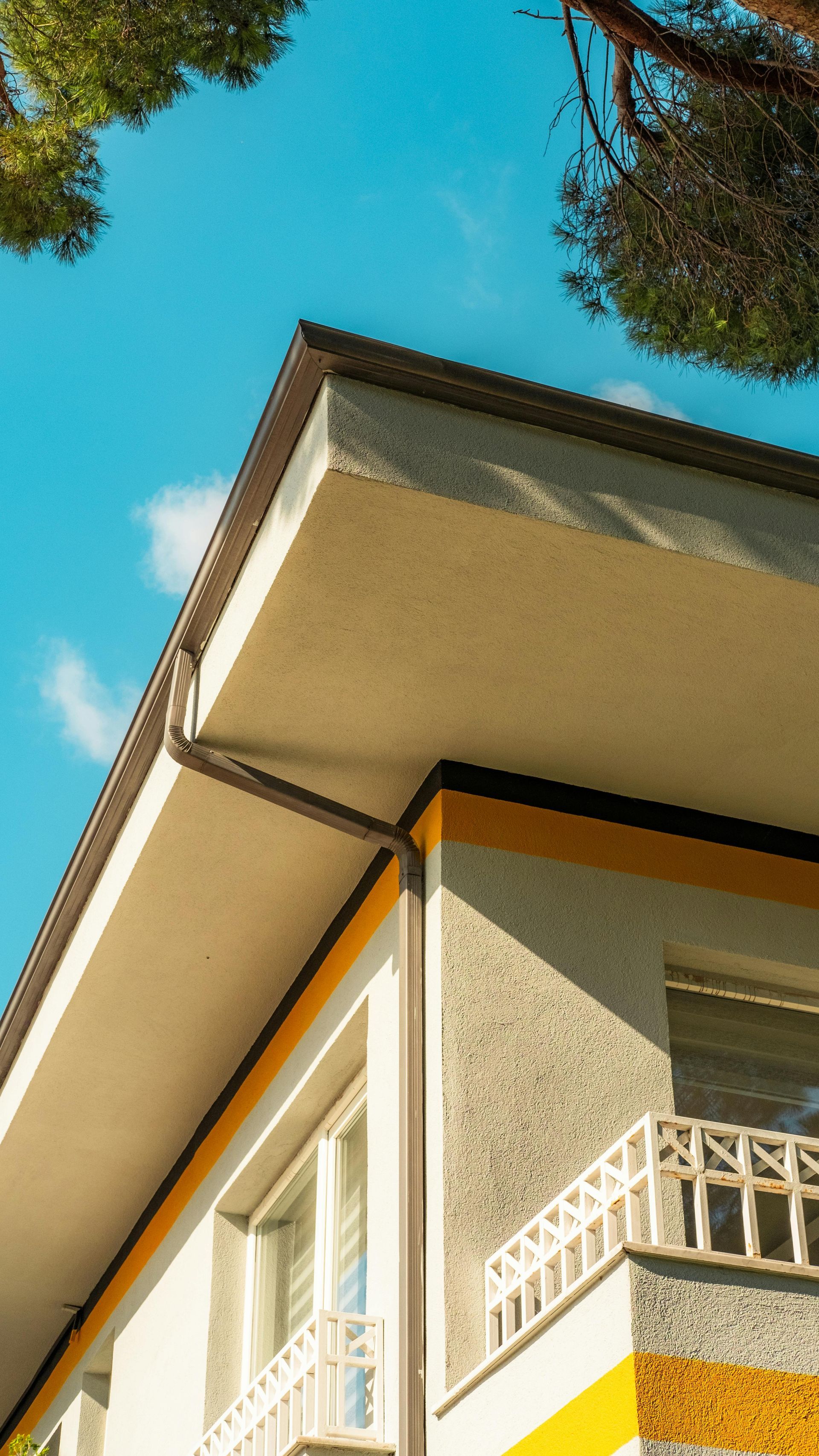 Low-angle view of a beige house corner with white balcony railings against a blue sky and tree branches