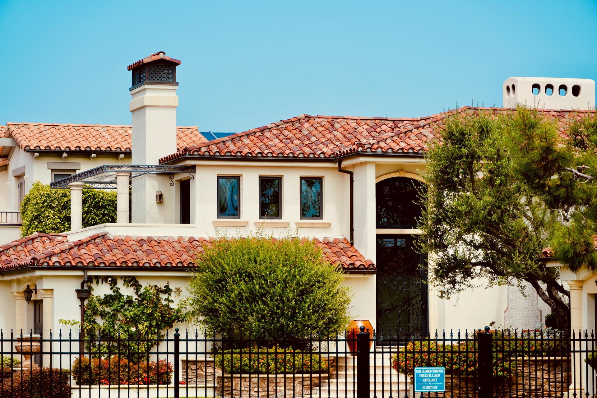 Two-story white house with red tile roof and black fence, surrounded by trees and shrubs