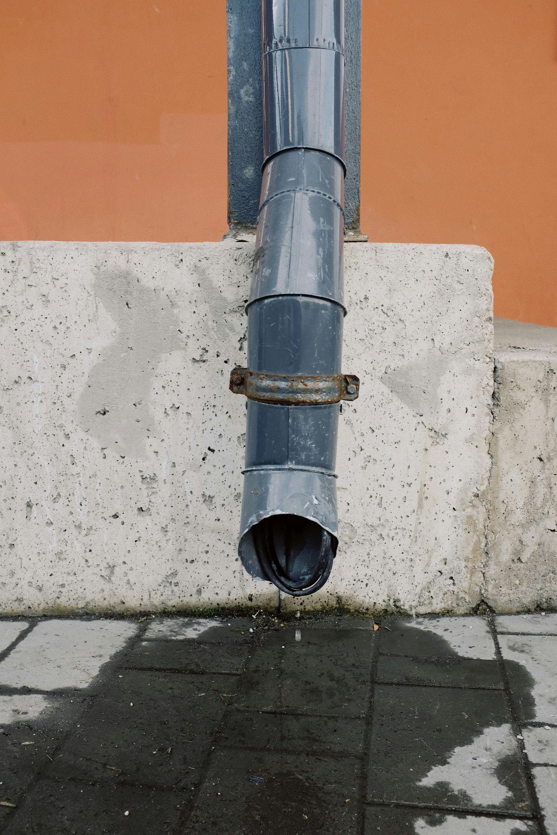 Metal downspout draining onto wet pavement beside a white wall and orange building.