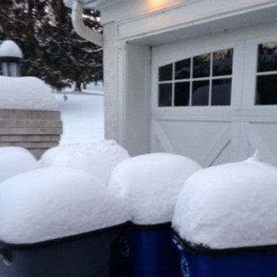 Snow piled on three blue trash bins beside a garage in winter.