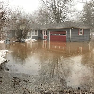 Flooded driveway in front of a gray house with a red garage door and snow along the edges