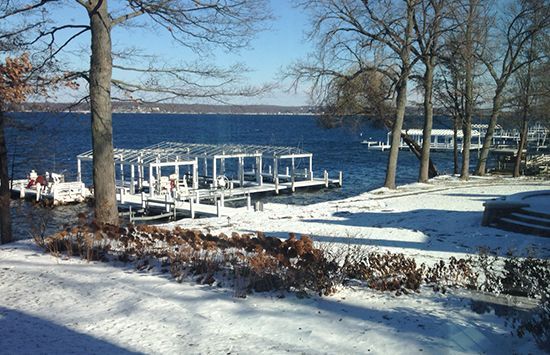 Snowy lakeside dock with bare trees and blue water under a clear sky