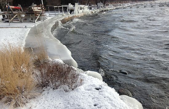Snowy lakeside path beside rough dark water, with dry reeds and shoreline erosion.