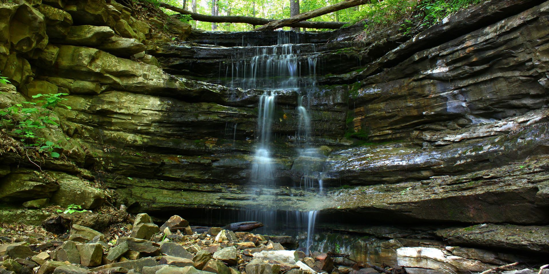 A thin, wispy waterfall cascades down a tiered, mossy rock cliff in a shaded, forested setting.