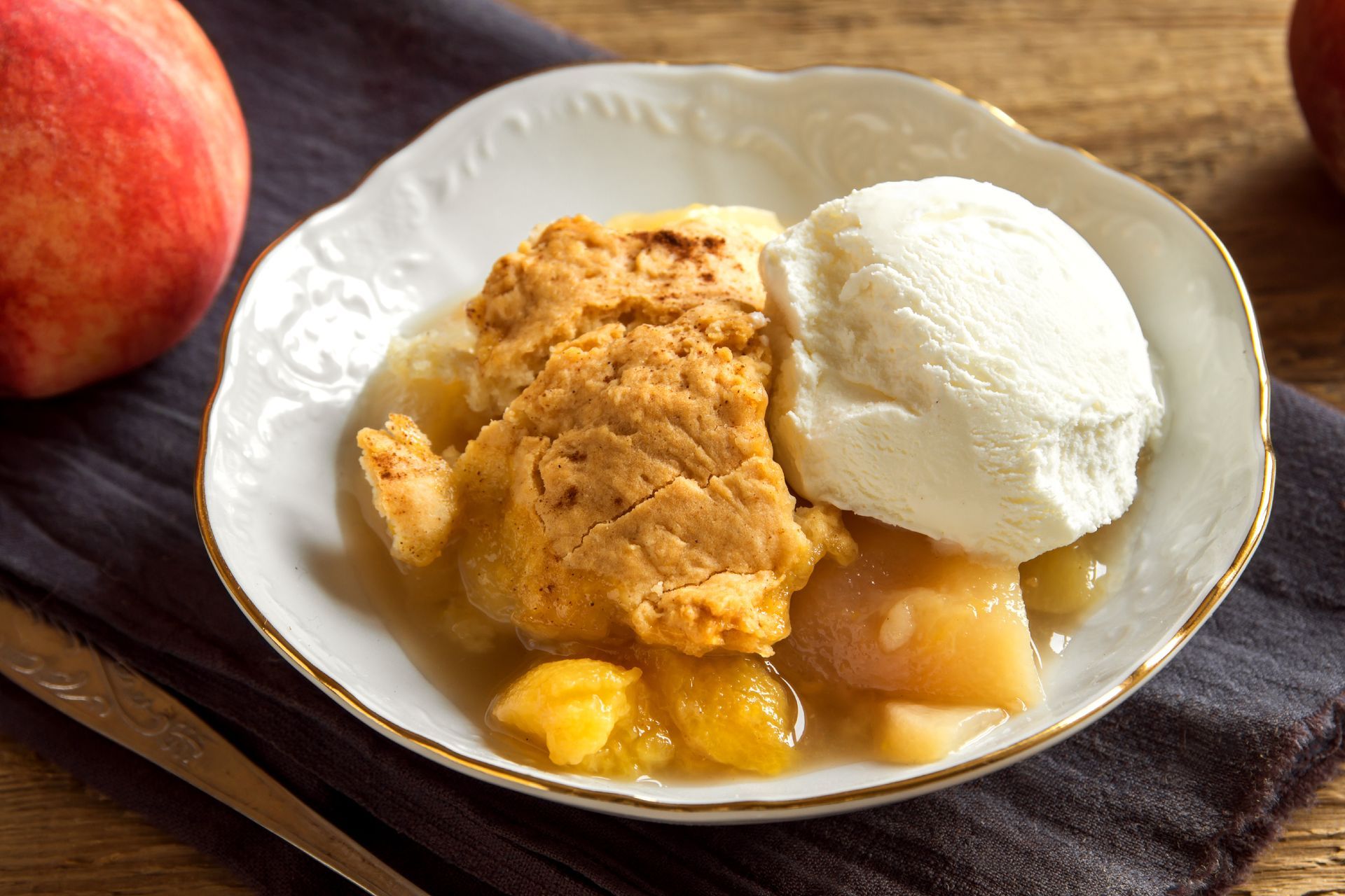 A bowl of peach cobbler topped with a scoop of vanilla ice cream, set on a dark cloth next to fresh peaches.