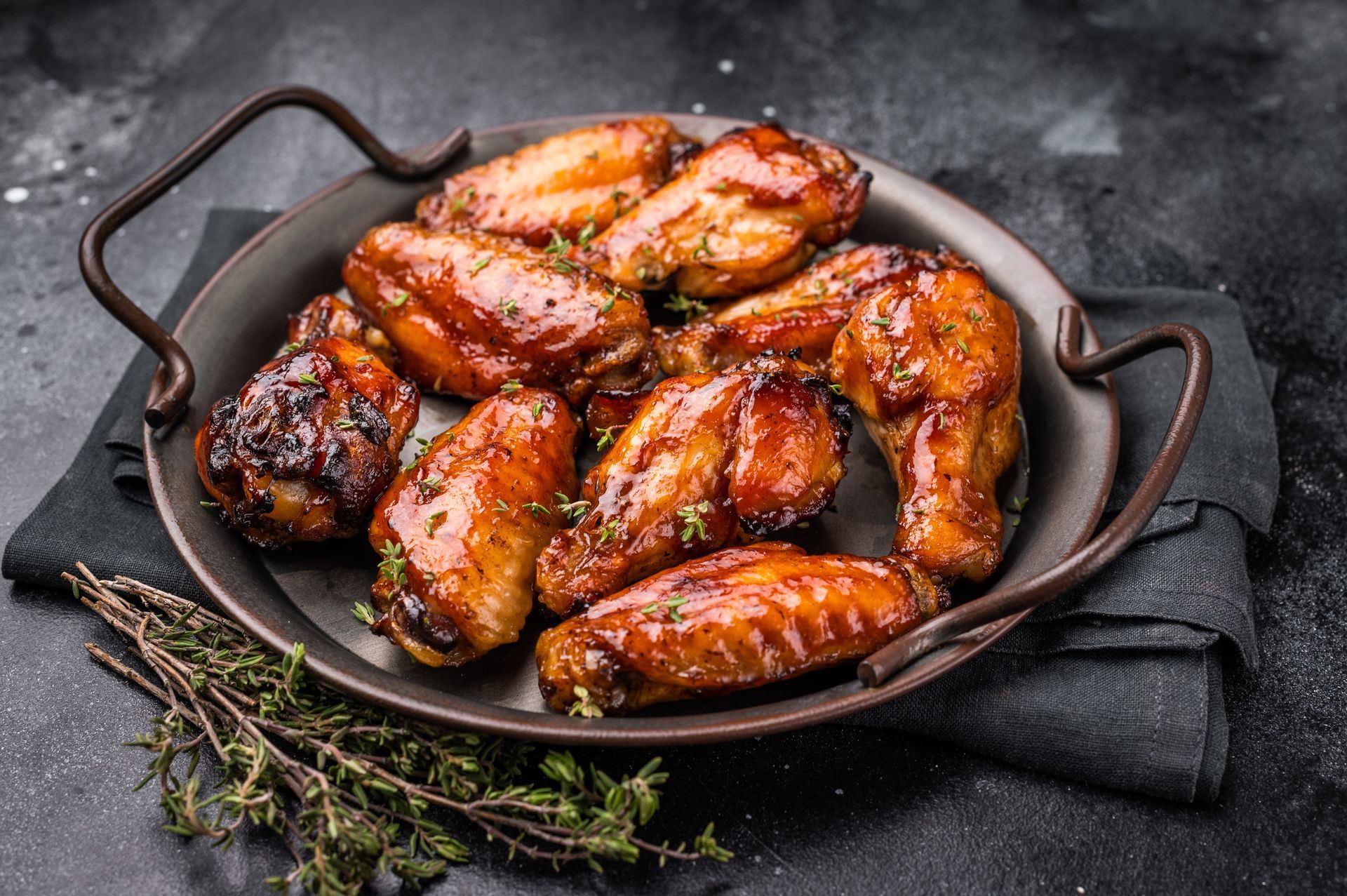 Glazed chicken wings in a dark metal pan on a black napkin, with fresh thyme sprigs on a dark stone surface.