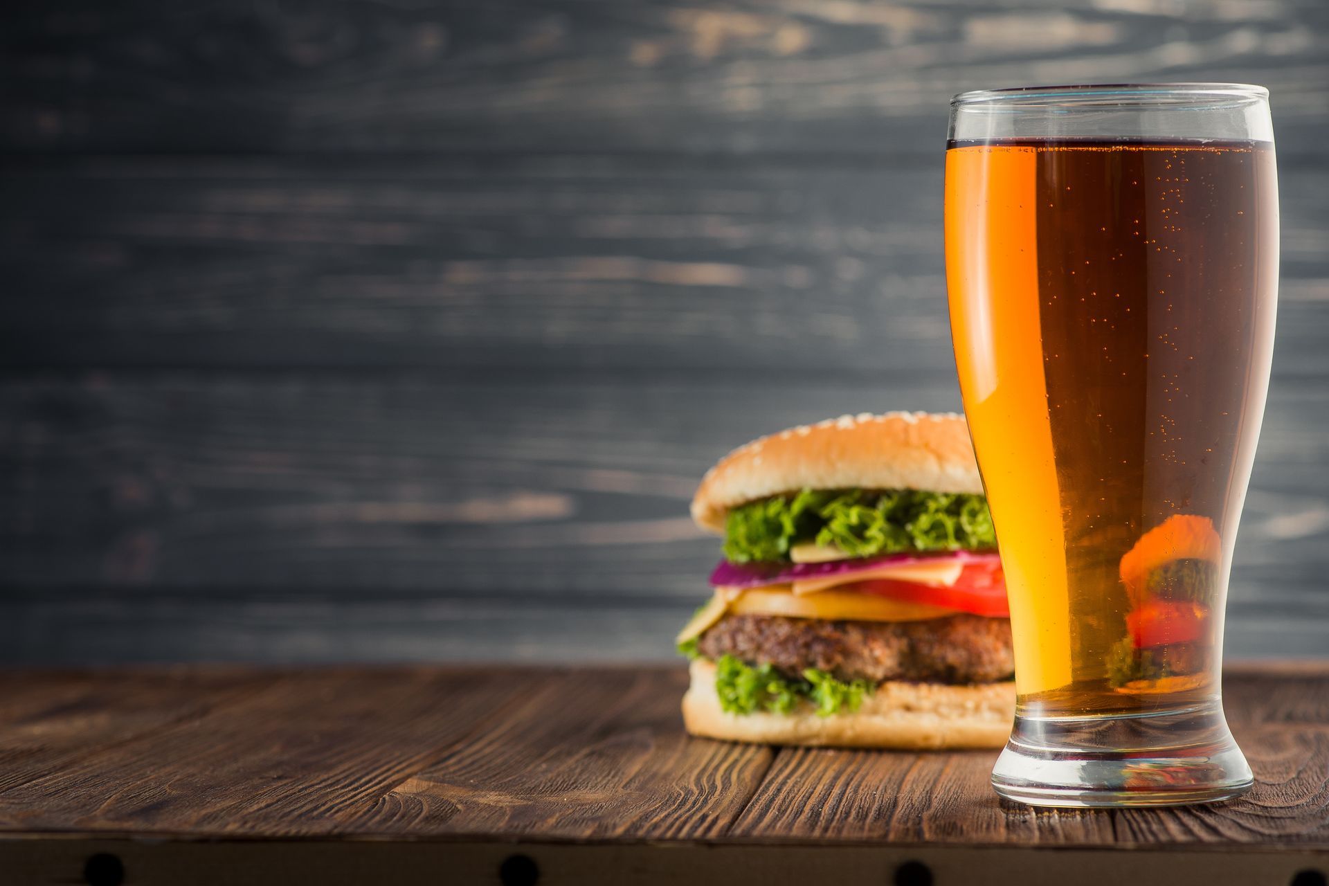 A glass of amber beer sits on a wooden table in front of a burger with fresh lettuce, tomato, and onion.