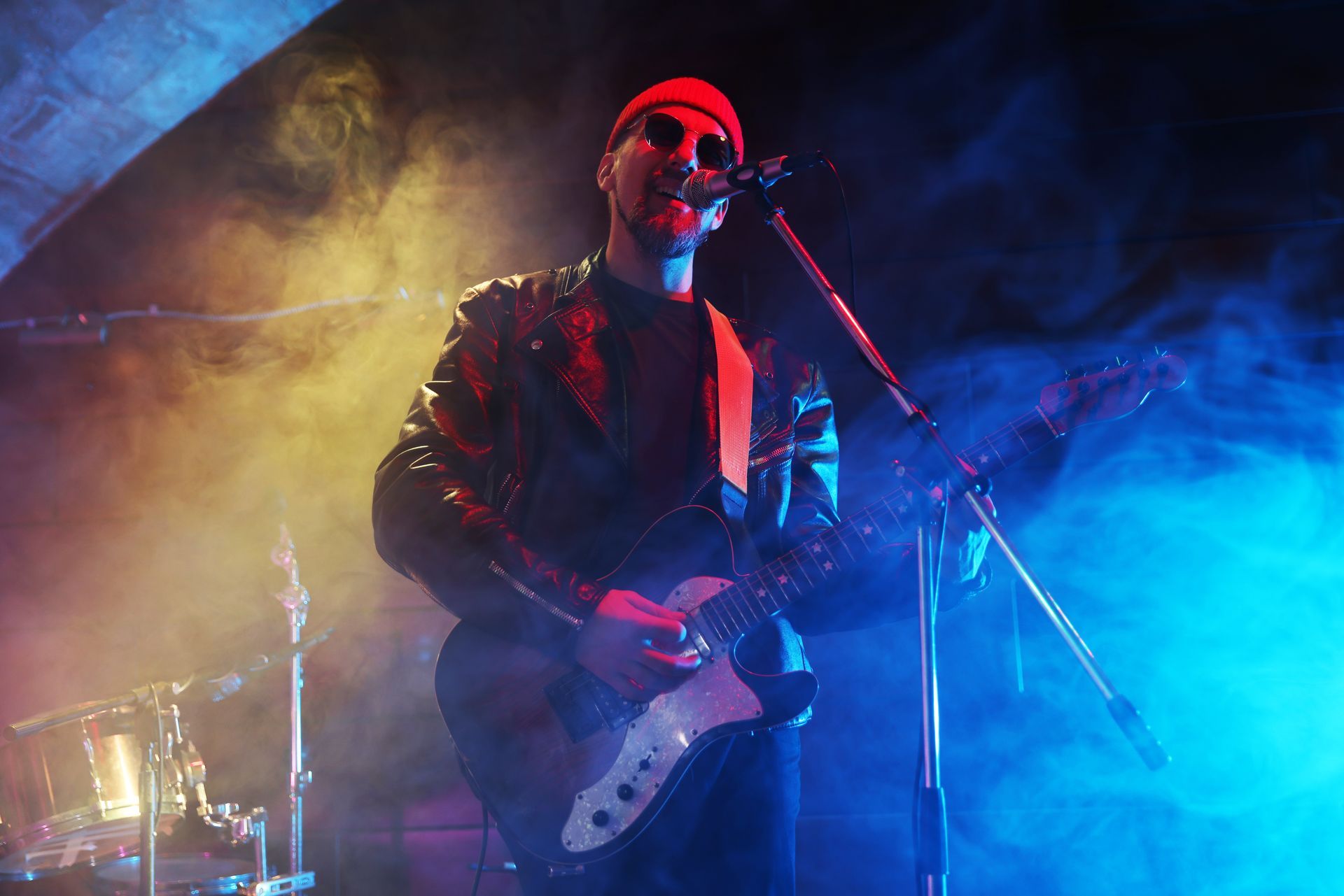 A musician in a red beanie and leather jacket plays an electric guitar under yellow and blue stage lights and smoke.