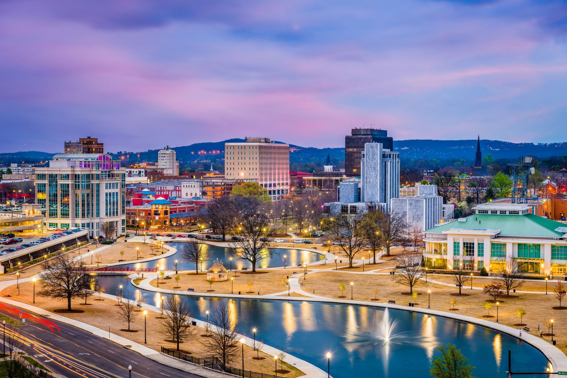 A scenic cityscape at dusk featuring a central pond, park trees, modern buildings, and a soft purple sunset.