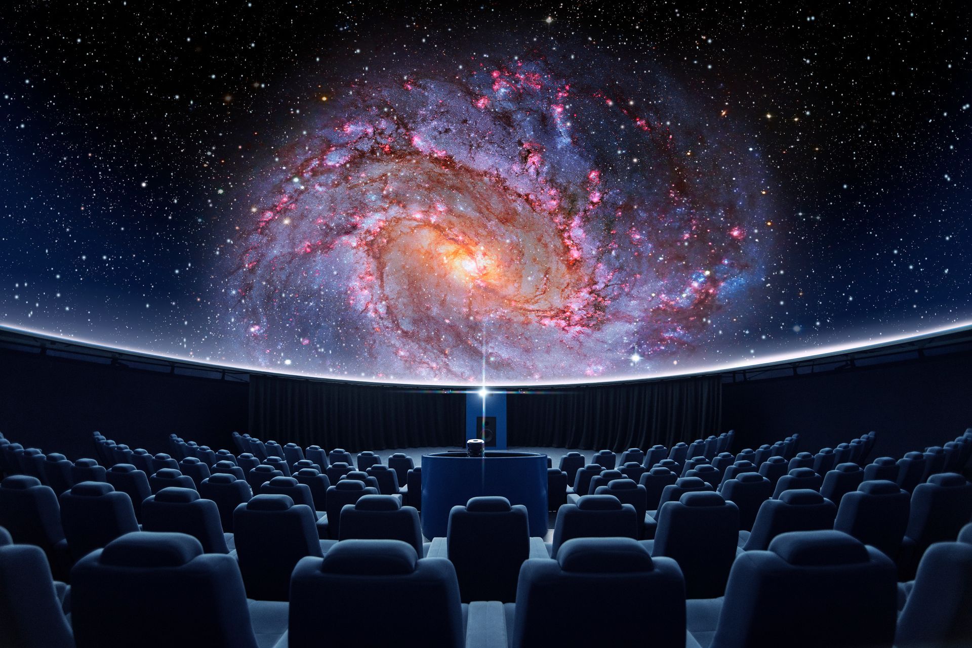 An empty planetarium theater with rows of dark seats facing a large dome screen displaying a bright, colorful galaxy.