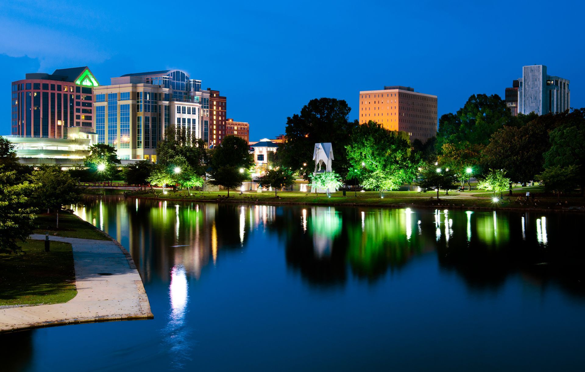 A city skyline at dusk with office buildings reflecting in a calm river, surrounded by illuminated trees and greenery