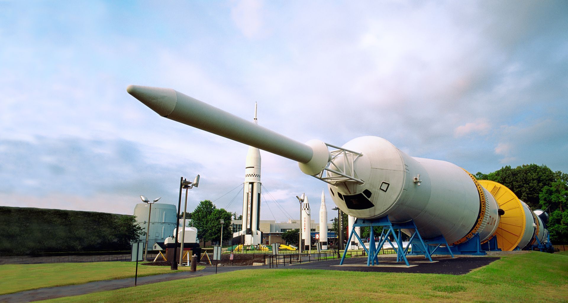 A large, horizontal space rocket exhibit displayed on a lawn at a park with a vertical rocket standing in the background.