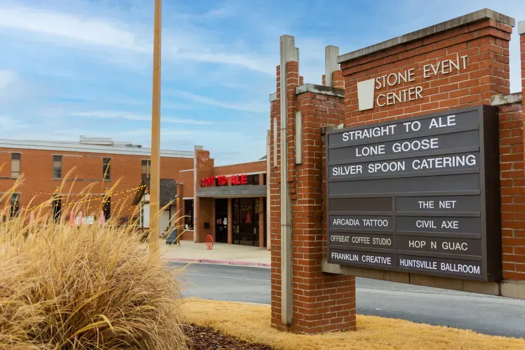 A brick sign for the Stone Event Center with event text, overlooking a brick building under a blue sky with tall grasses.