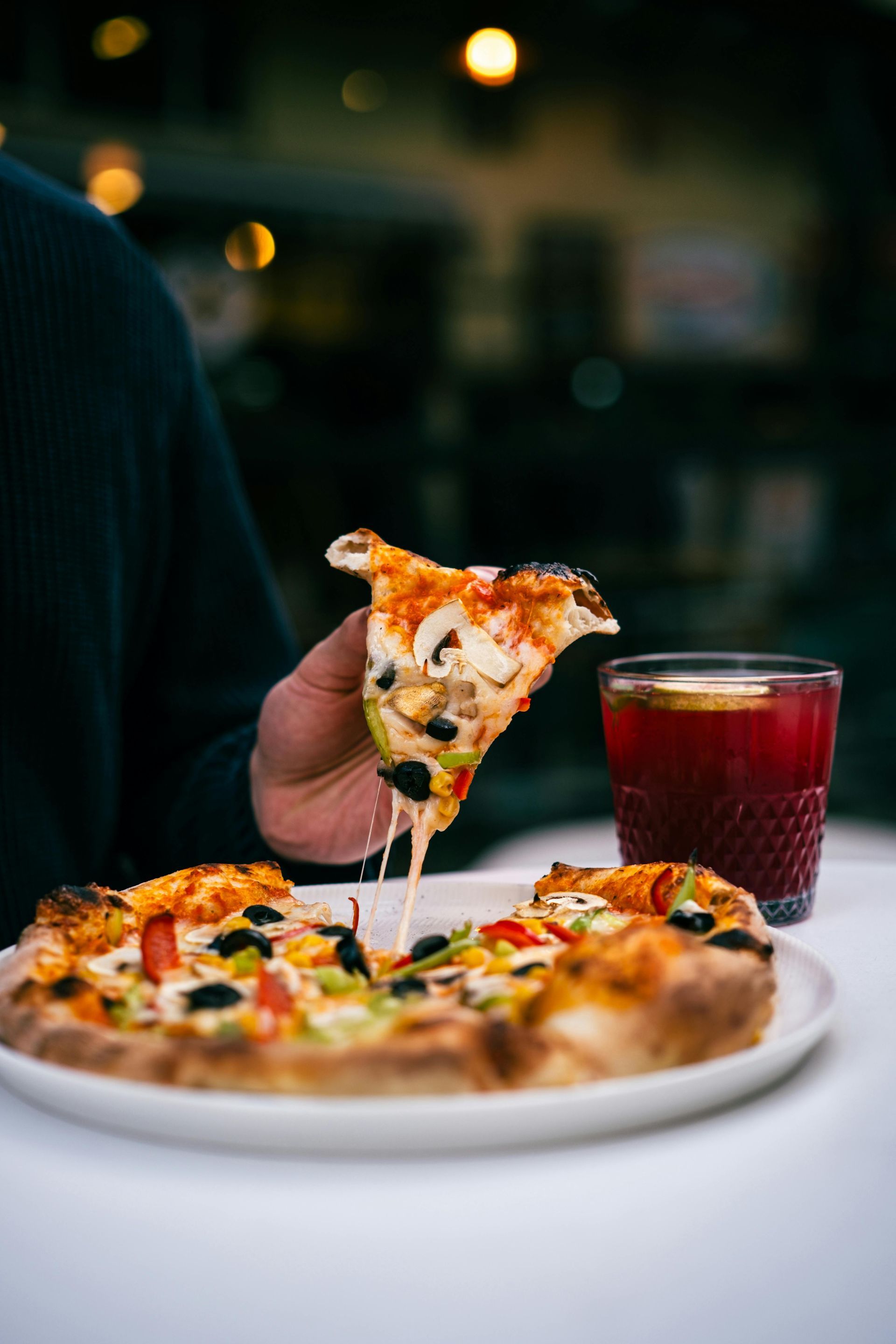 A hand holds a slice of vegetable pizza with melted cheese above a whole pizza on a plate next to a glass of red drink.