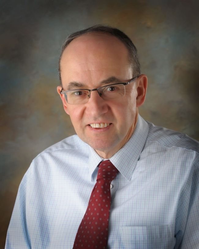 Portrait of a man in glasses, light blue shirt, and red tie against a blurred studio background