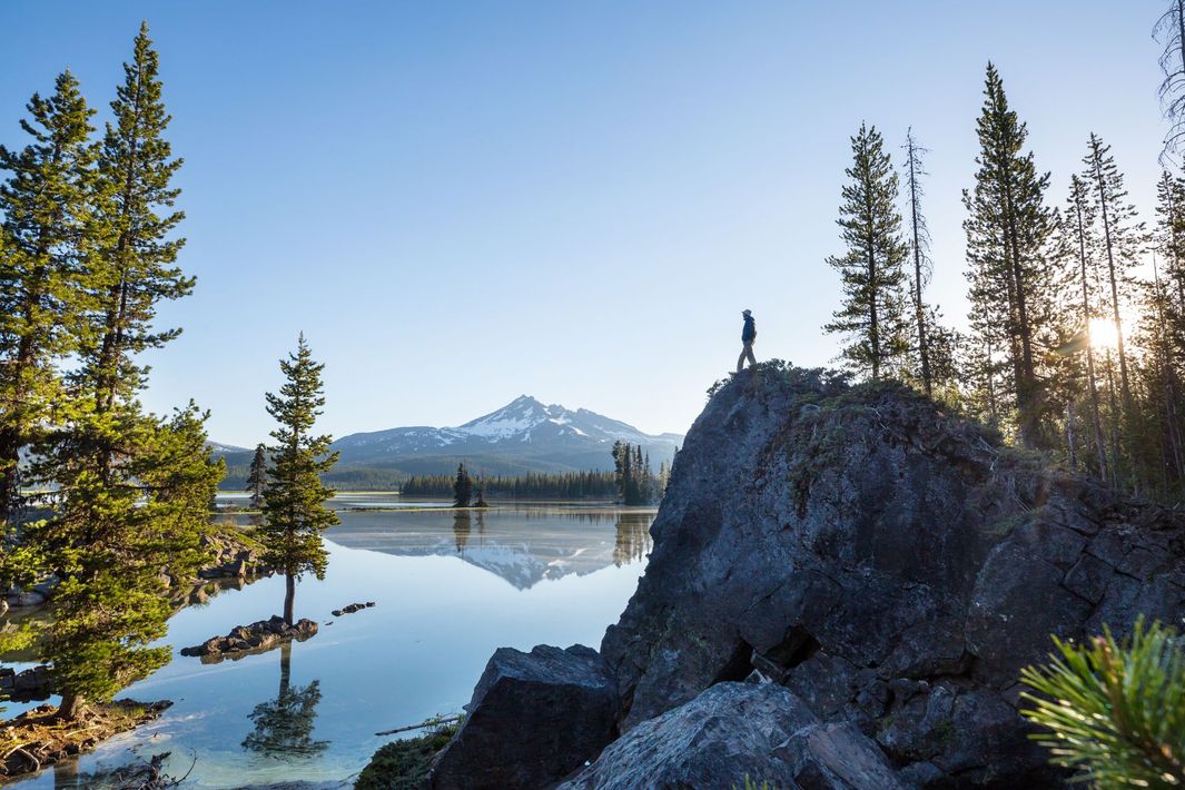 Snowy mountain reflected in a calm lake, with evergreen trees and rocky shore at sunrise.