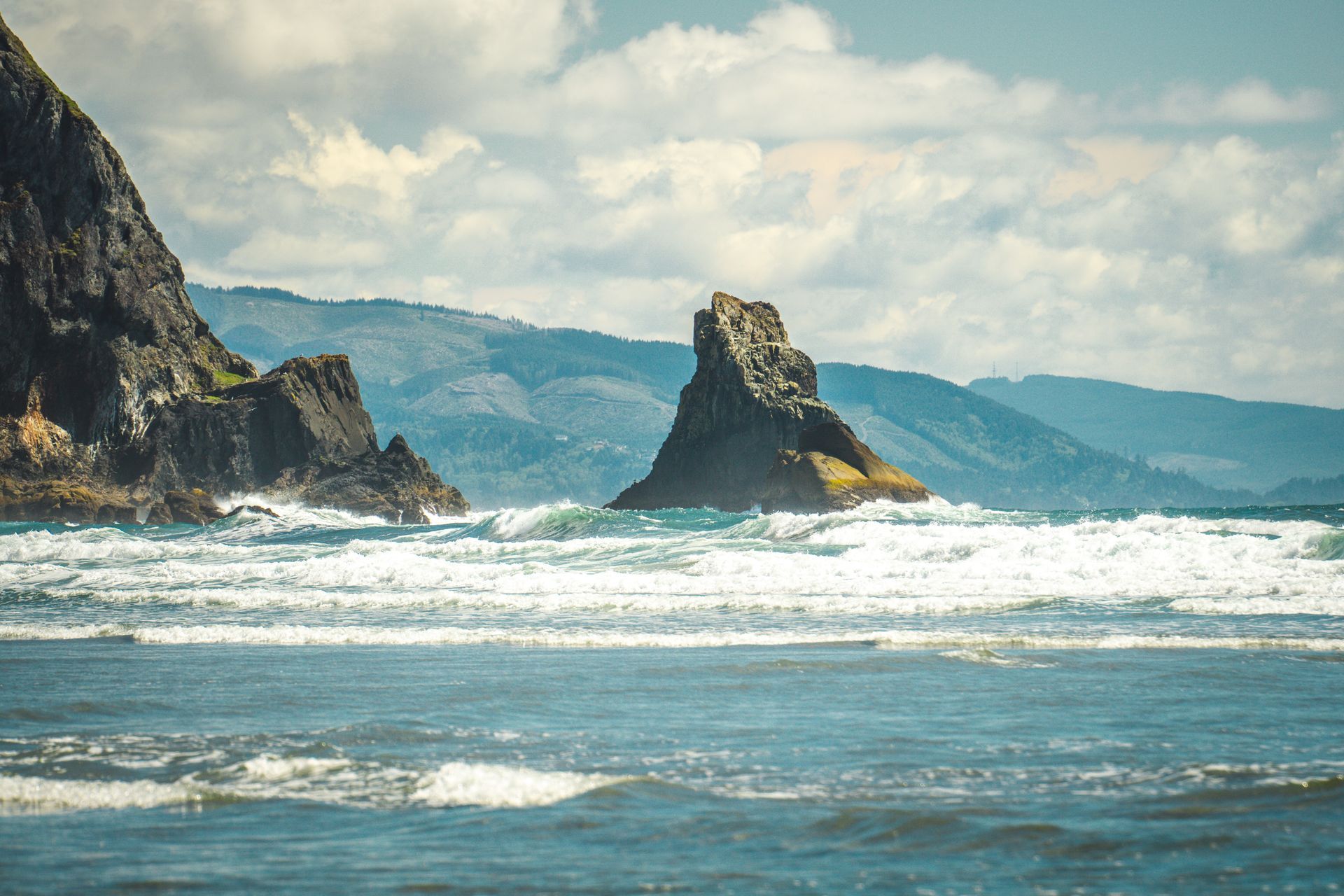 Rocky sea stack by a turbulent ocean, with cliffs and misty mountains under a cloudy sky