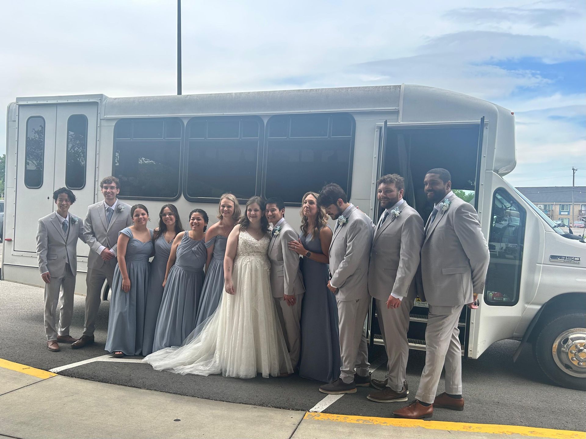 Bride, groom, and wedding party standing beside the white 15-passenger party bus