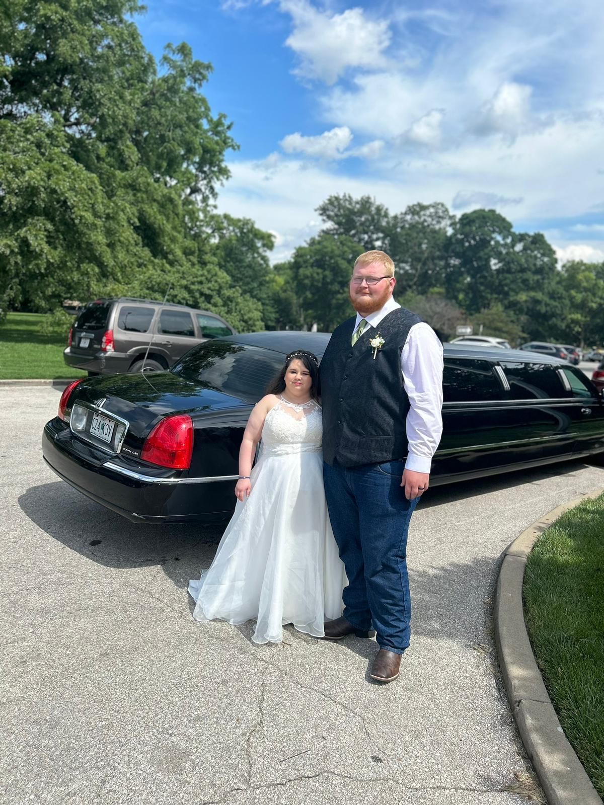Couple in formal attire standing beside a black limousine on a gravel drive with trees in the background