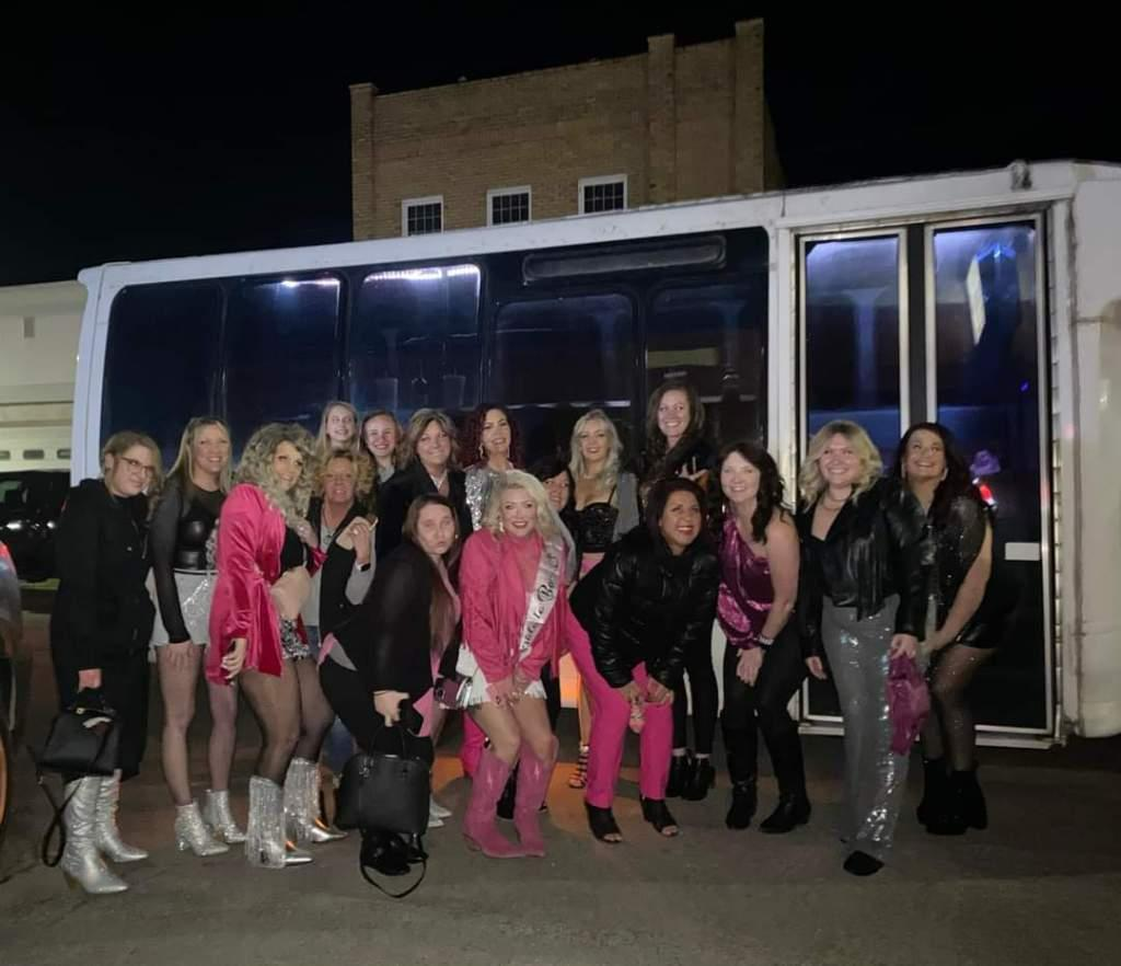 Group posing in front of a white bus at night, many wearing black and pink outfits.