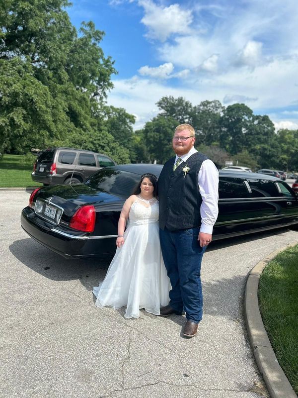 Couple posing outdoors beside a black limousine in a park driveway, both dressed formally.