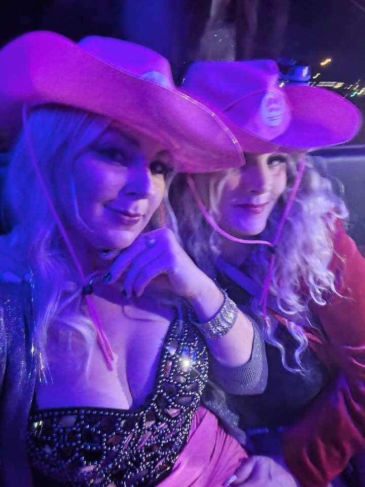 Two women in cowboy hats pose at a purple-lit event, wearing sparkly outfits and smiling.