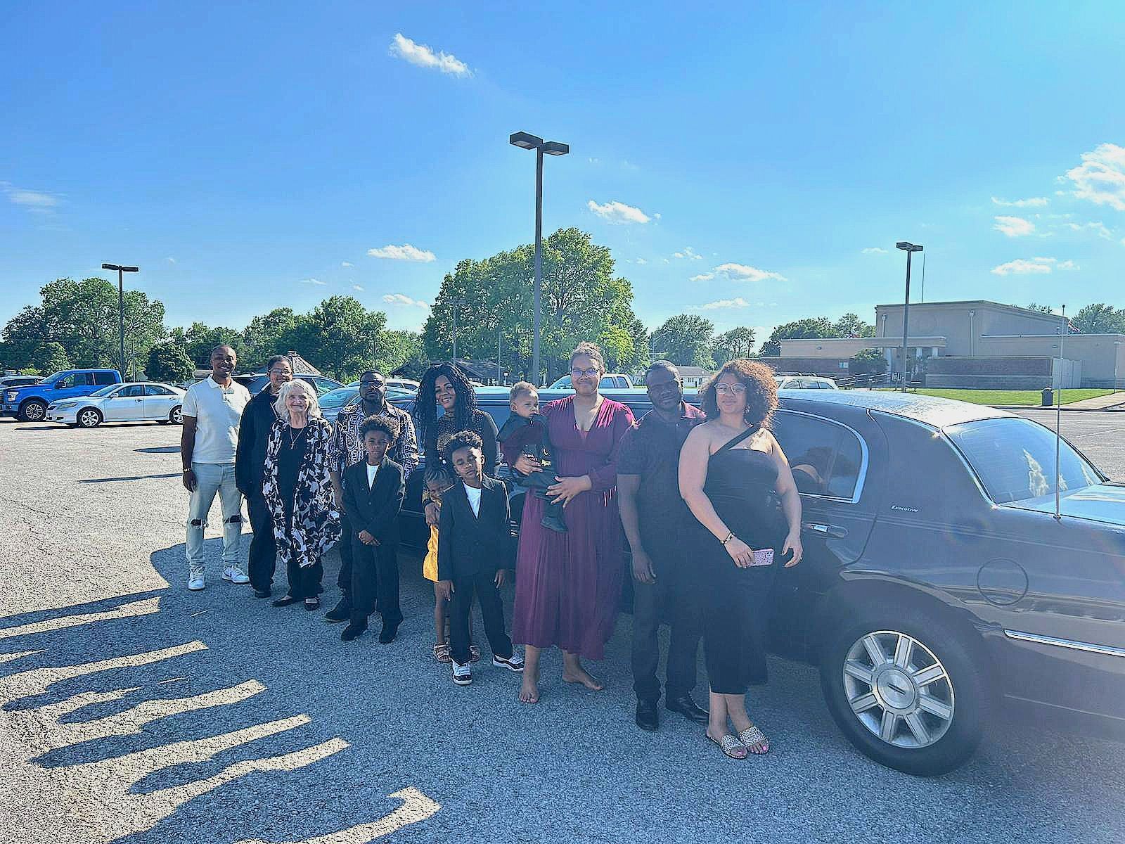 Group dressed for a formal event stands beside a black car in a sunny parking lot.