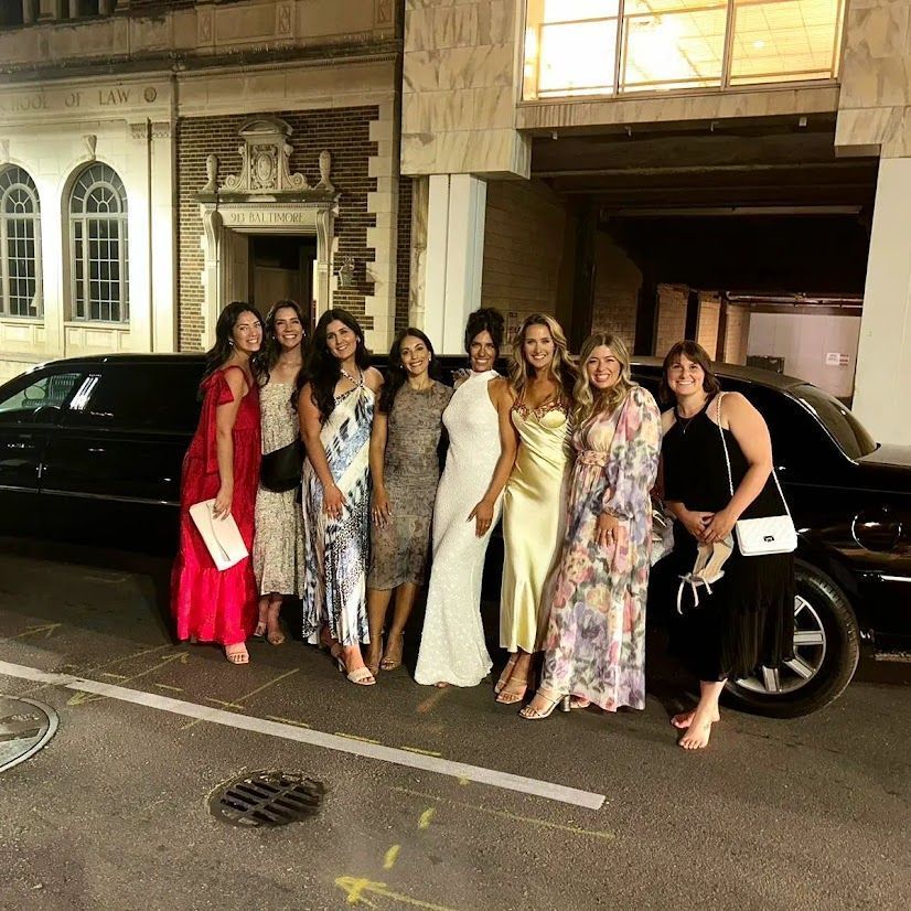 Group of women posing on a city street at night beside parked cars, dressed in colorful formal outfits.